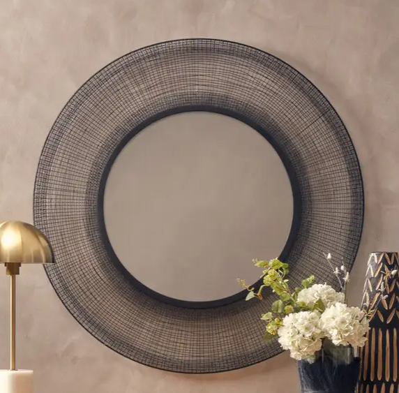 Black wireframe mirror on a brown wall with lamp and flowers beside it