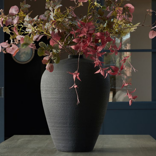 A large matt black vase with autumnal flowers in it, on a table